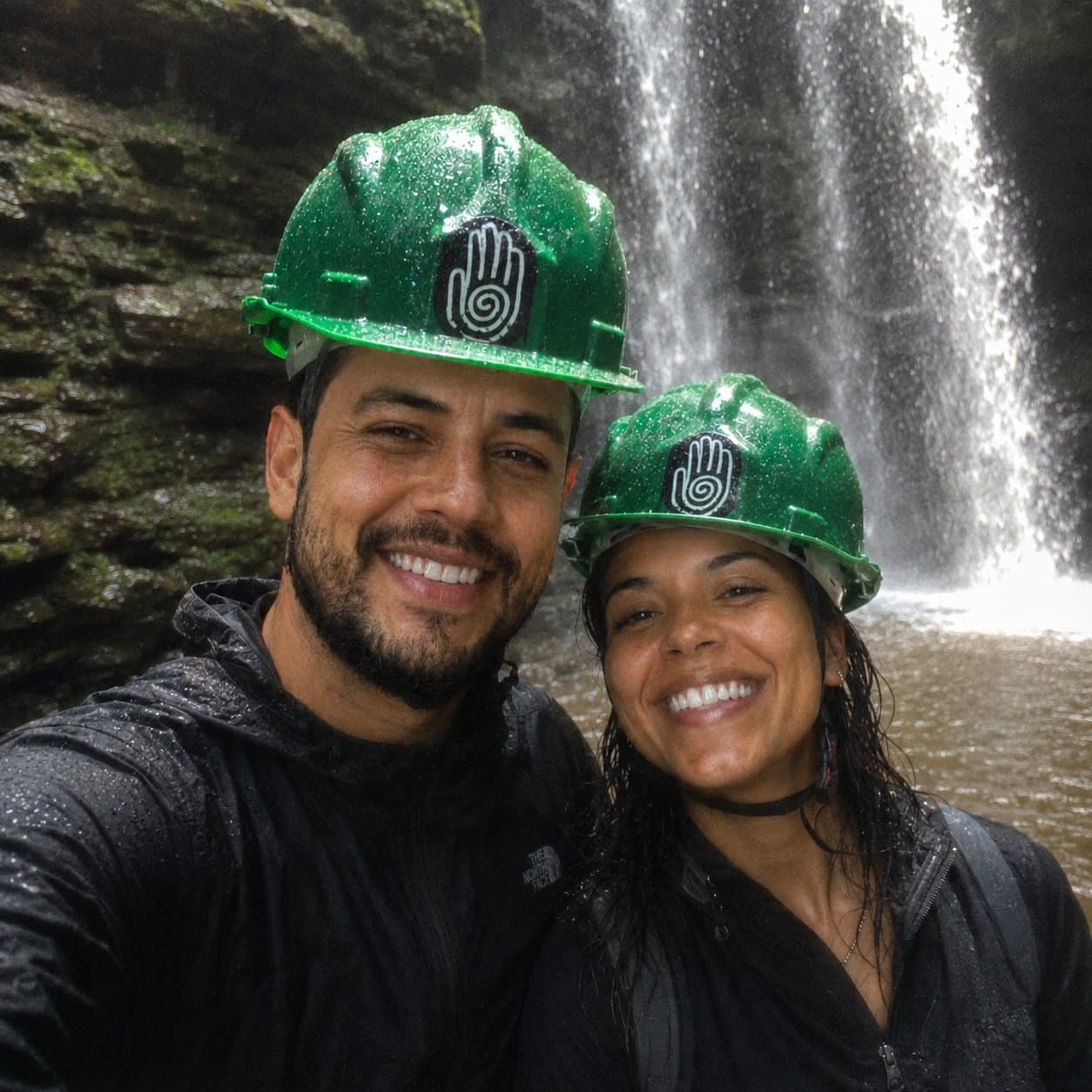 Matheus e Luana de capacete verde em frente à cachoeira